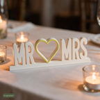 Deko-Schriftzug Mr & Mrs auf Hochzeitstafel umgeben von brennenden Teelichtern, KI-generiert
